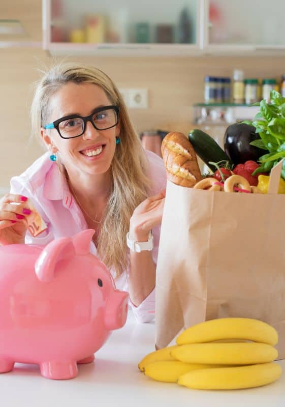 Woman in the kitchen with full grocery bag and piggy bank in the counter.