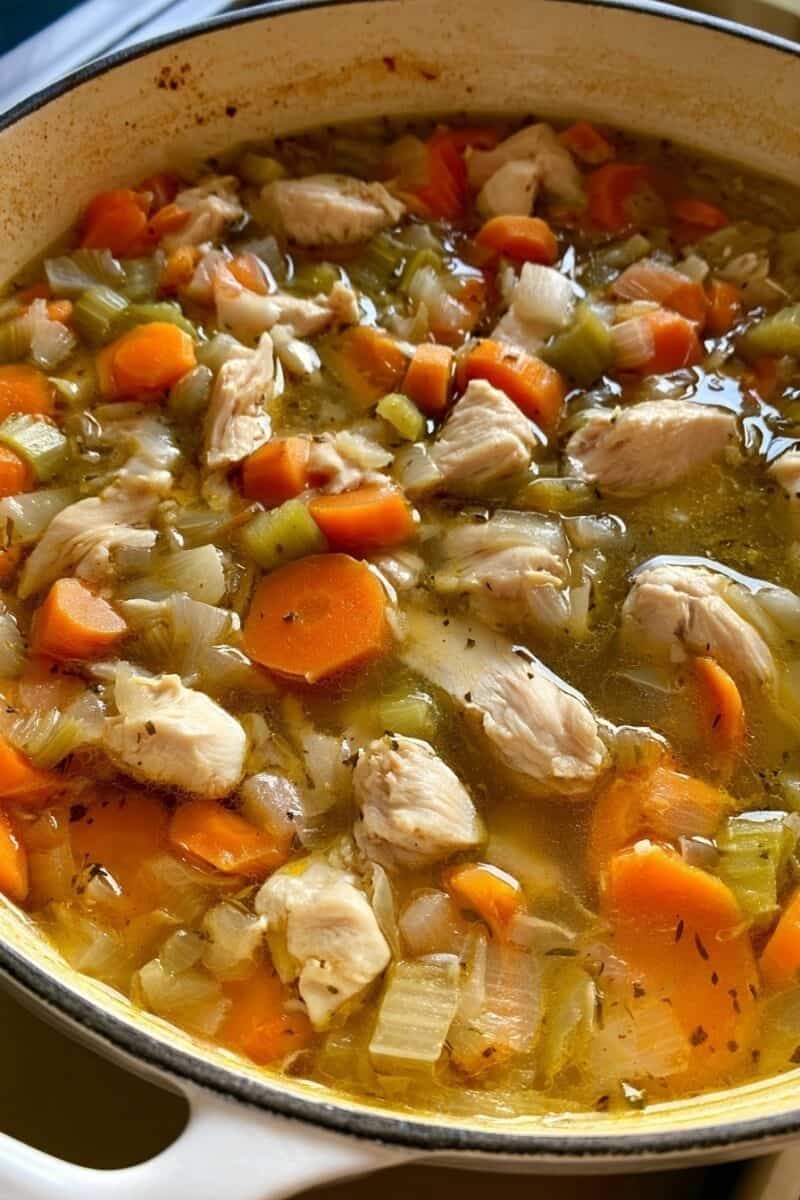 Close-up of bite-sized chicken breast pieces, sliced carrots, diced celery, and onions simmering in golden chicken broth inside a white Dutch oven before the noodles are added.
