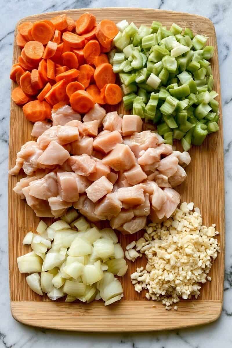 Overhead view of a wooden cutting board with sliced carrots, chopped celery, raw chicken breast cubes, diced yellow onion, and minced garlic arranged in separate sections on a marble countertop.