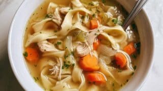 Overhead view of a white bowl filled with homemade chicken noodle soup, featuring wide egg noodles, shredded chicken, sliced carrots, celery, and herbs in golden broth, with a silver spoon resting inside.