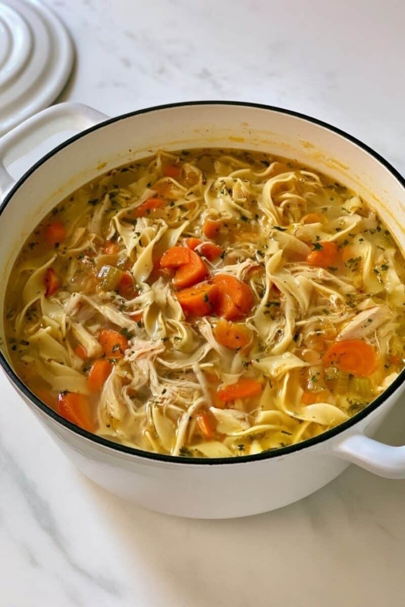 White Dutch oven filled with homemade chicken noodle soup, showing shredded chicken, wide egg noodles, sliced carrots, celery, and herbs in golden broth on a marble countertop.