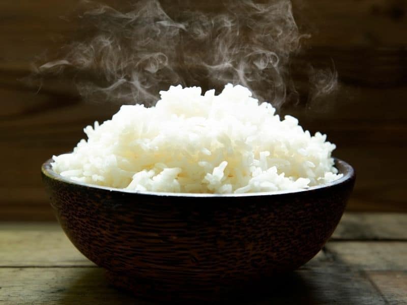 Side view of a black bowl of steaming rice on a dark background.