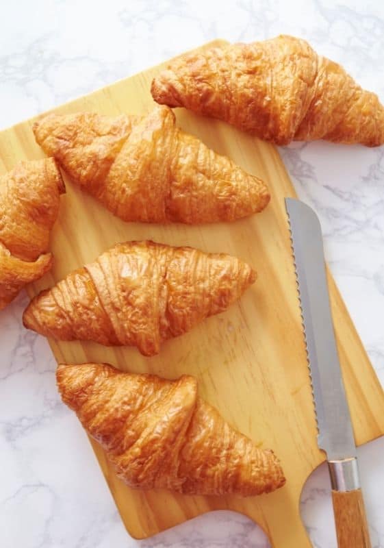 Five Baked croissants on a wooden cutting board.