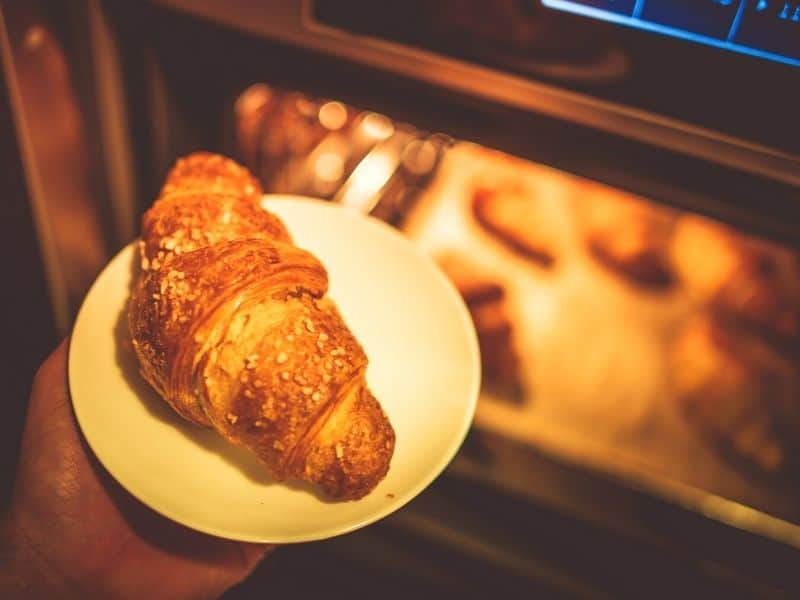 Oven with croissants being baked and one croissant in a plate.