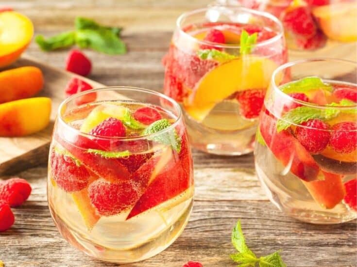 Three delicious glasses of Sparkling Wine Sangria garnished with peaches, raspberries and mint leaves.