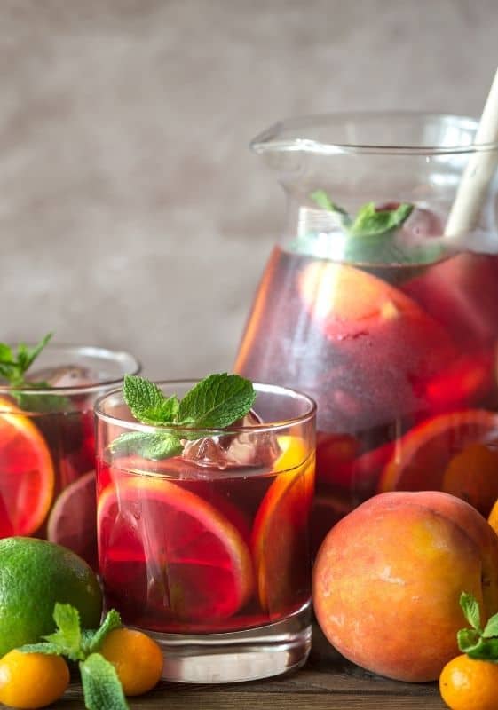 Side view of fruity Spanish sangria pitcher and two glasses garnished with fruits in a gray background.