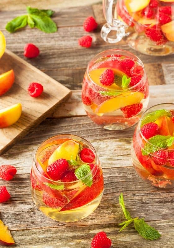 Top view of three glasses of Sparkling Wine Sangria with peaches and raspberries with a cutting board on the background with fruits on it.