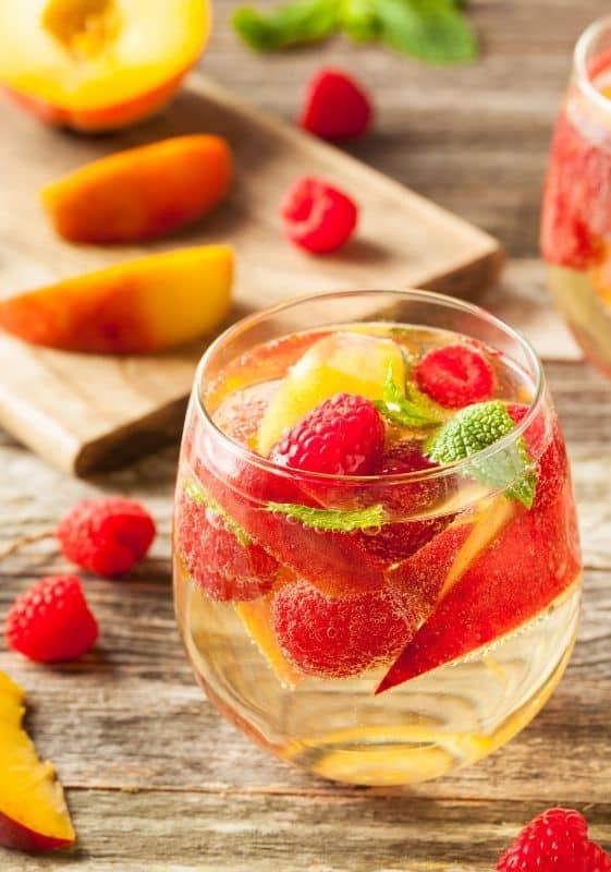 Side view of a glass of Sparkling Wine Sangria with peaches and raspberries with a cutting board on the background with fruits on it.
