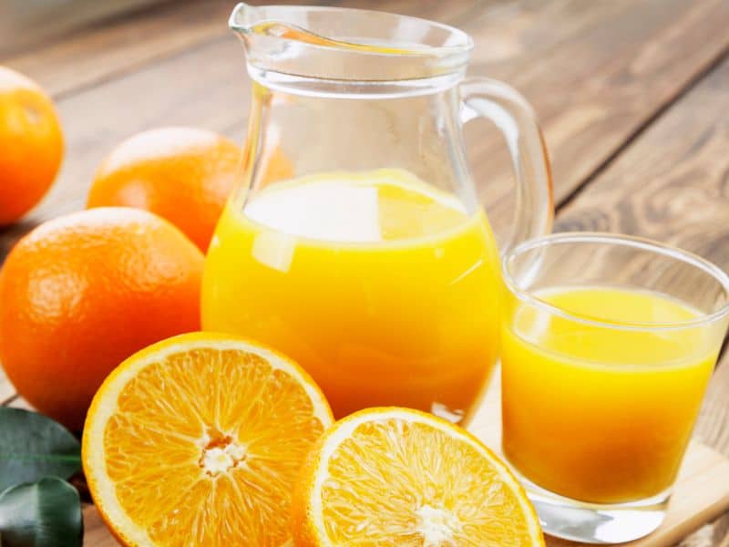 Pitcher and glass of orange juice with whole and half oranges around it on a wooden table.