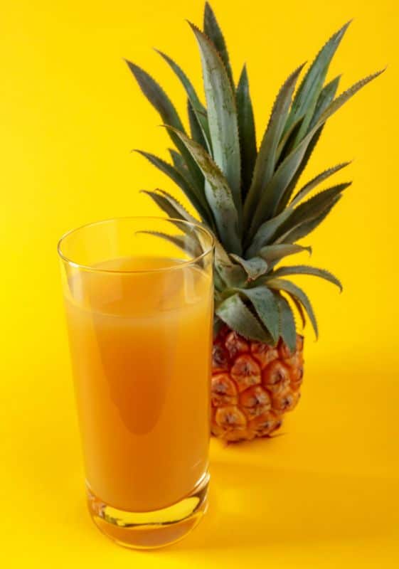 A glass of pineapple juice with a whole pineapple on a yellow background.