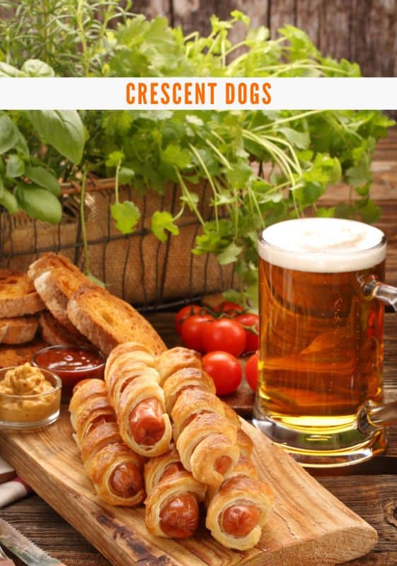 View of Crescent hot dogs stacked up on a rectangular wooden cutting board with a pint of beer on the side.