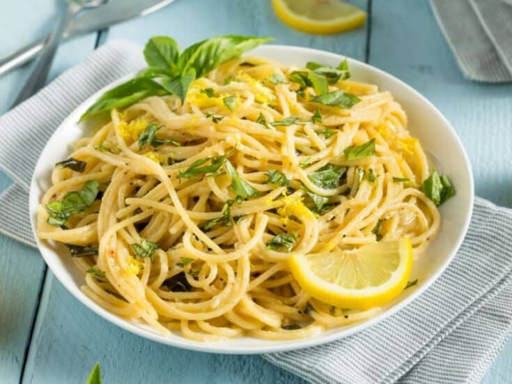 A white plate with butter lemon pasta sauce garnished with basil and lemon slice.
