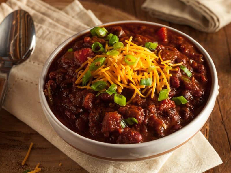 The best classic chili recipe in a white with one red stripe bowl topped with cheese and scallions.