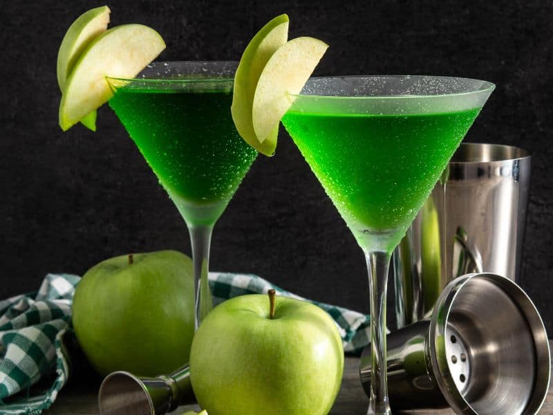 A visually stunning. Appletini cocktail, served in a martini glass, with a perfectly sliced green apple resting on the rim. The vibrant green color of the cocktail is highlighted, enticing the viewer's taste buds.