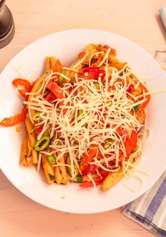 Rasta Pasta in a white plate and a fork. A beautifully presented Rasta Pasta dish on a white plate, artistically garnished with parmesan cheese. The creamy sauce gently clings to the pasta, adorned with colorful bell peppers, offering a delightful and appetizing sight.