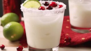 A glass of White Christmas Margarita on a festive table, rimmed with sanding sugar and garnished with a lime slice and cranberries, surrounded by holiday decorations.