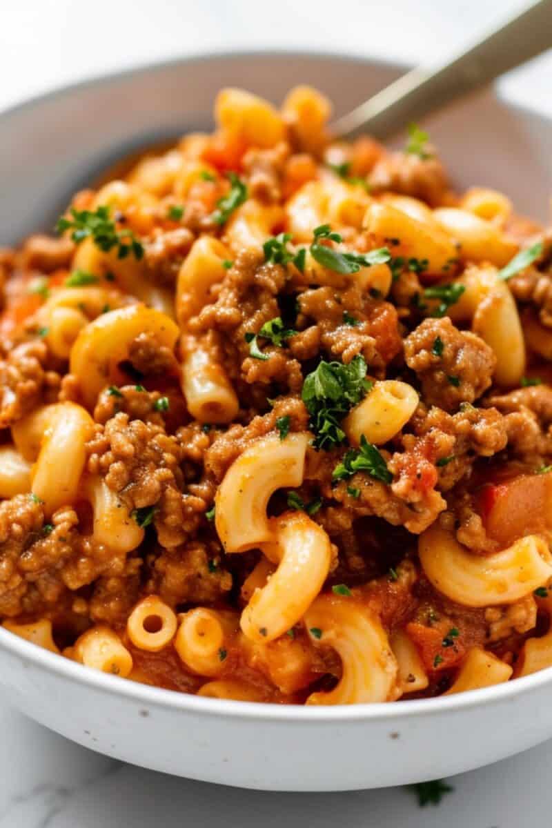 Image of a white bowl filled with homemade Beefaroni, featuring tender macaroni and savory ground beef enveloped in a rich, tomato-based sauce, garnished with a sprinkle of fresh herbs.