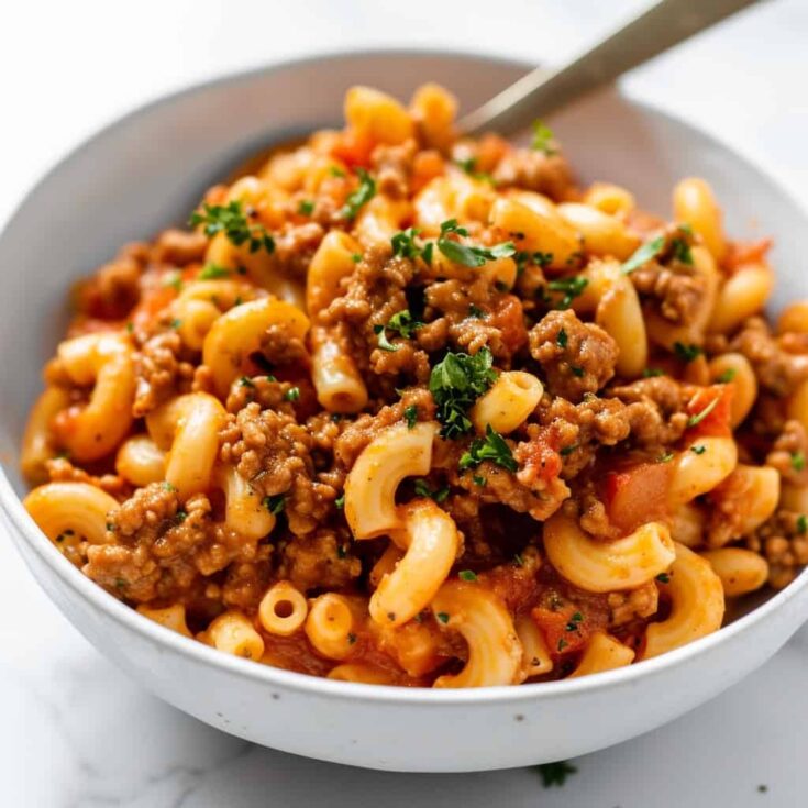 Image of a hearty serving of Beefaroni in a white bowl, showcasing the rich, meaty sauce with ground beef, macaroni pasta, and hints of herbs, garnished with a sprinkle of grated cheese on top. The dish is set on a rustic wooden table, indicating a home-cooked, comforting meal.