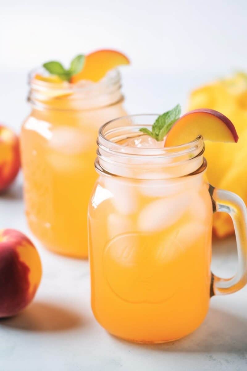 Two mason jars of peach lemonade on an outdoor picnic table, each jar brimming with cold, peach-infused lemonade, ice cubes, and garnished with fresh peach slices, conveying a sense of a refreshing summer drink.