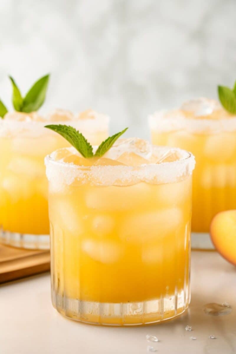 A close-up of Big Batch Peach Margaritas in clear glasses, showcasing the golden hue of the drink, with a backdrop of peaches and mint leaves.
