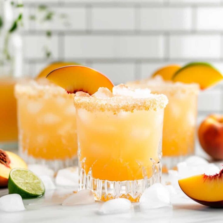 Glasses of Peach Margaritas on a white background, garnished with lime wheels and peach slices, embodying the refreshing taste of summer.