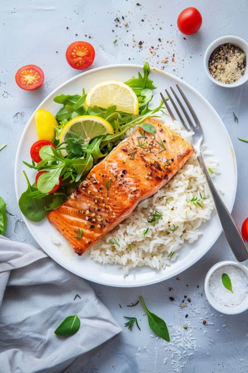 Overhead view of Pan Seared Salmon with lemon slices on top, alongside cilantro rice and a vibrant side salad on a white plate, set on a white table.