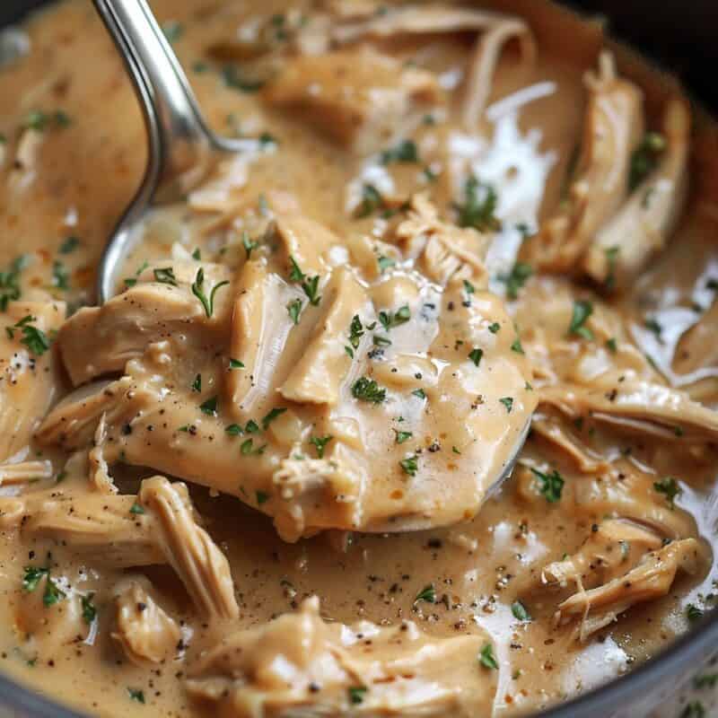 A spoon lifts tender, juicy chicken thighs drenched in rich gravy from a crockpot, highlighting the dish's succulence.