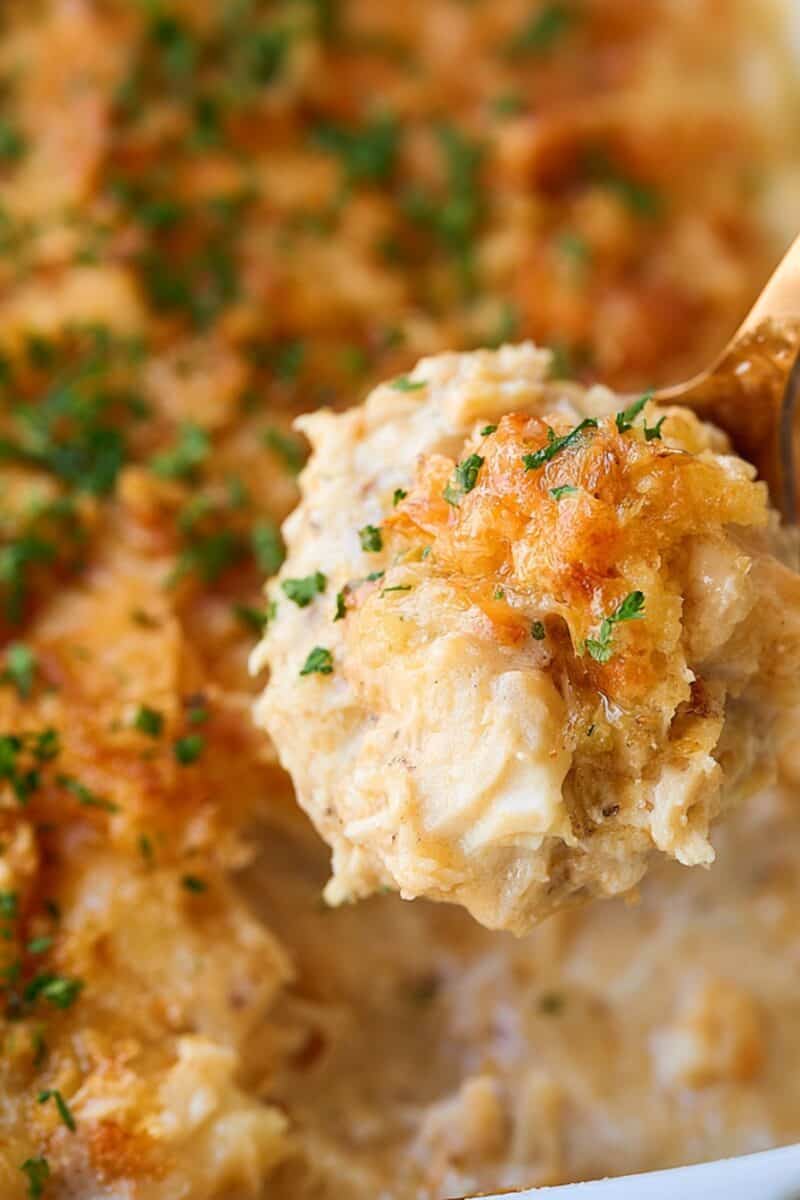 Close-up of creamy Million Dollar Crockpot Chicken on a serving spoon, topped with a golden crunchy topping and fresh herbs, with the chicken mixture in the slow cooker below.