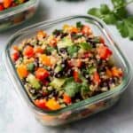 A vibrant Quinoa and Black Bean Salad, meticulously layered in a clear glass storage container. The bottom layer showcases fluffy, perfectly cooked quinoa, followed by a generous portion of glossy black beans. Bright red bell pepper and crisp cucumber add a burst of color and texture, while finely chopped fresh cilantro sprinkles the top.