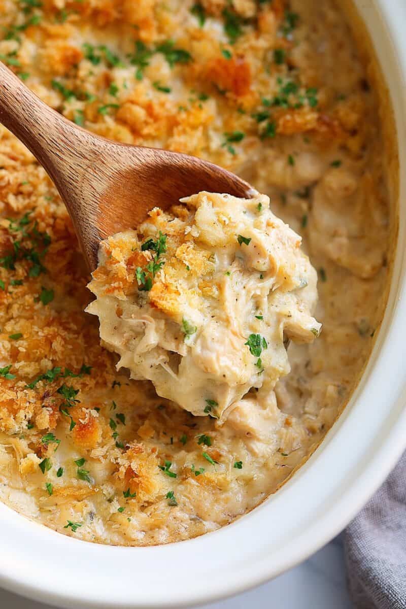 Wooden spoon lifting a serving of creamy Crockpot Million Dollar Chicken with tender shredded chicken, a golden crispy topping, and fresh chopped herbs from a white slow cooker.
