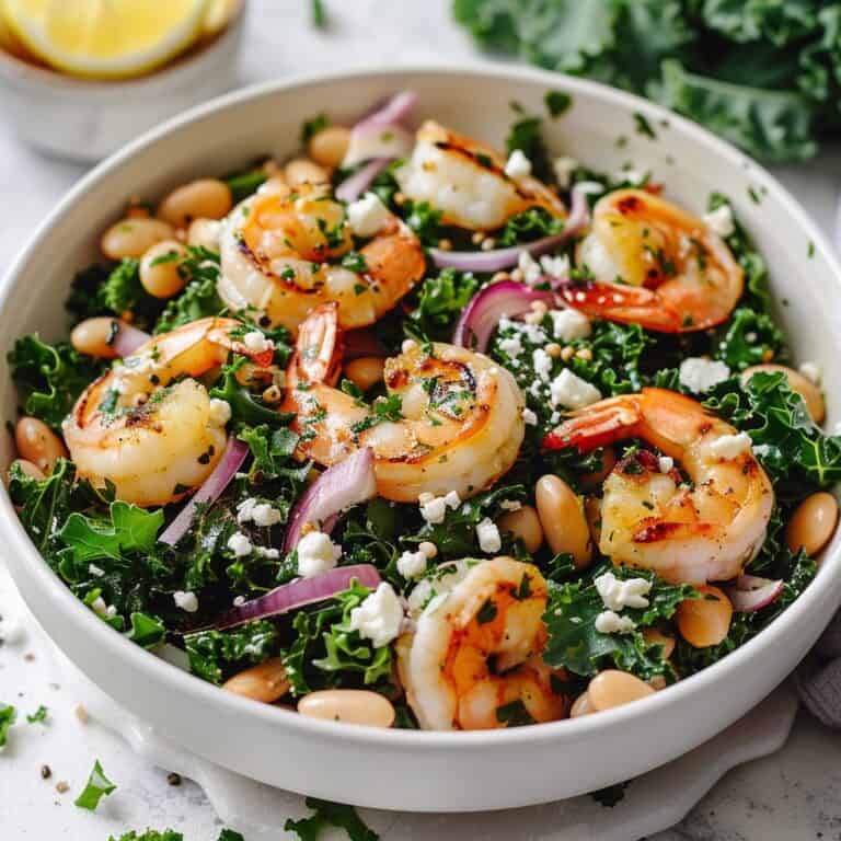 A vibrant bowl of Kale White Bean Salad topped with golden grilled shrimp, sprinkled with seeds and crumbled feta, all drizzled with lemony dressing.