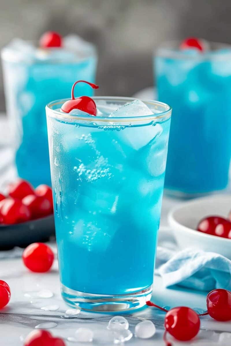 Three glasses filled with vibrant blue lemonade are arranged on a gray background, with one glass in super close focus, showcasing the fizz and the Maraschino cherry garnish in vivid detail, while the other two glasses softly blur into the backdrop, creating a striking contrast.