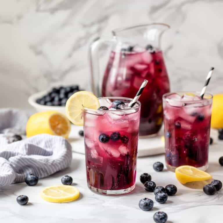 On a white counter, clear pitcher and glasses filled with blueberry lemonade, garnished blueberries, offering an inviting, refreshing scene.