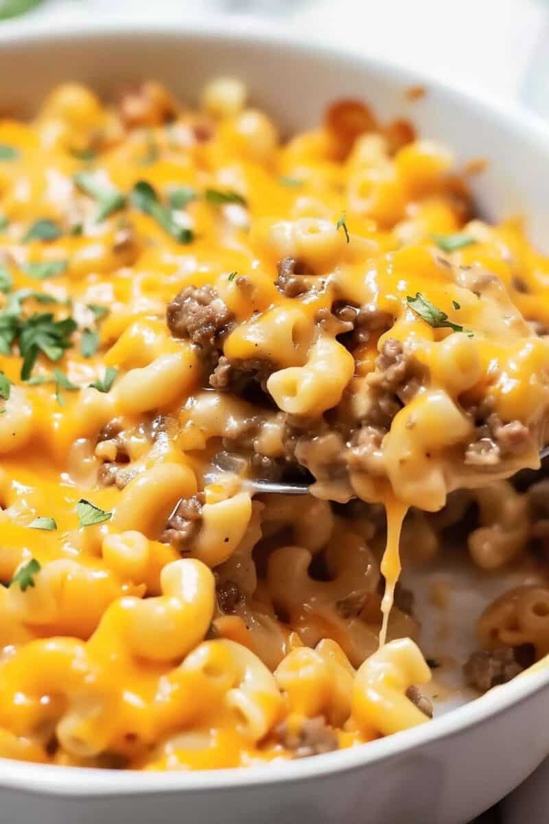 
A golden-brown Hamburger Mac and Cheese Casserole in a baking dish, with a spoon lifting a serving to reveal a stretchy, cheesy pull.