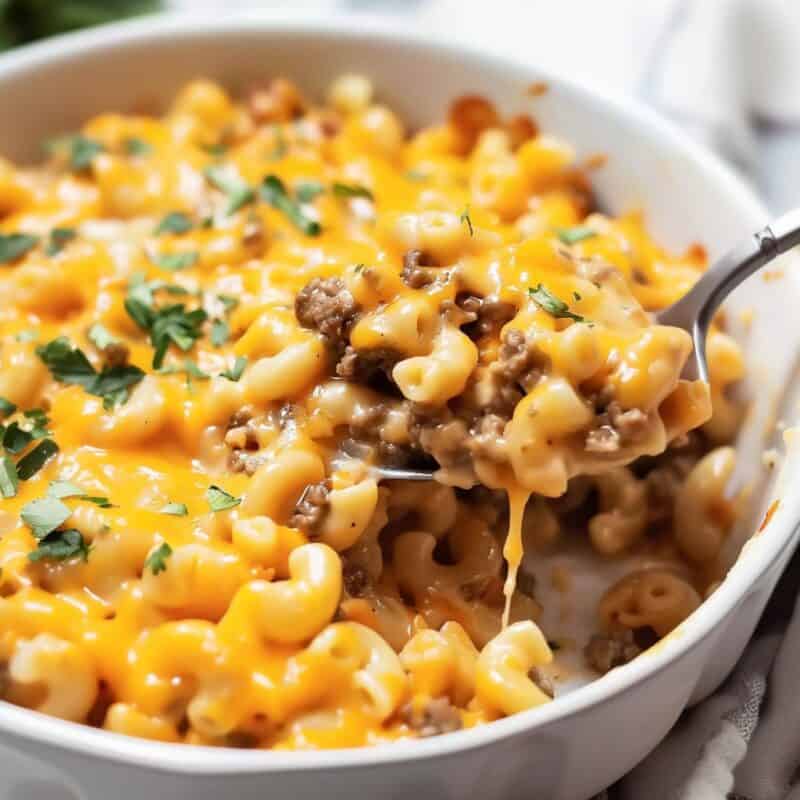 A golden-brown Hamburger Mac and Cheese Casserole in a baking dish, with a spoon lifting a serving to reveal a stretchy, cheesy pull.