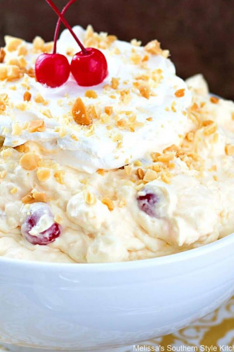 A close-up of a creamy layered dessert topped with whipped cream, a sprinkle of nuts, and a pair of maraschino cherries in a white bowl, suggesting a rich and decadent texture.