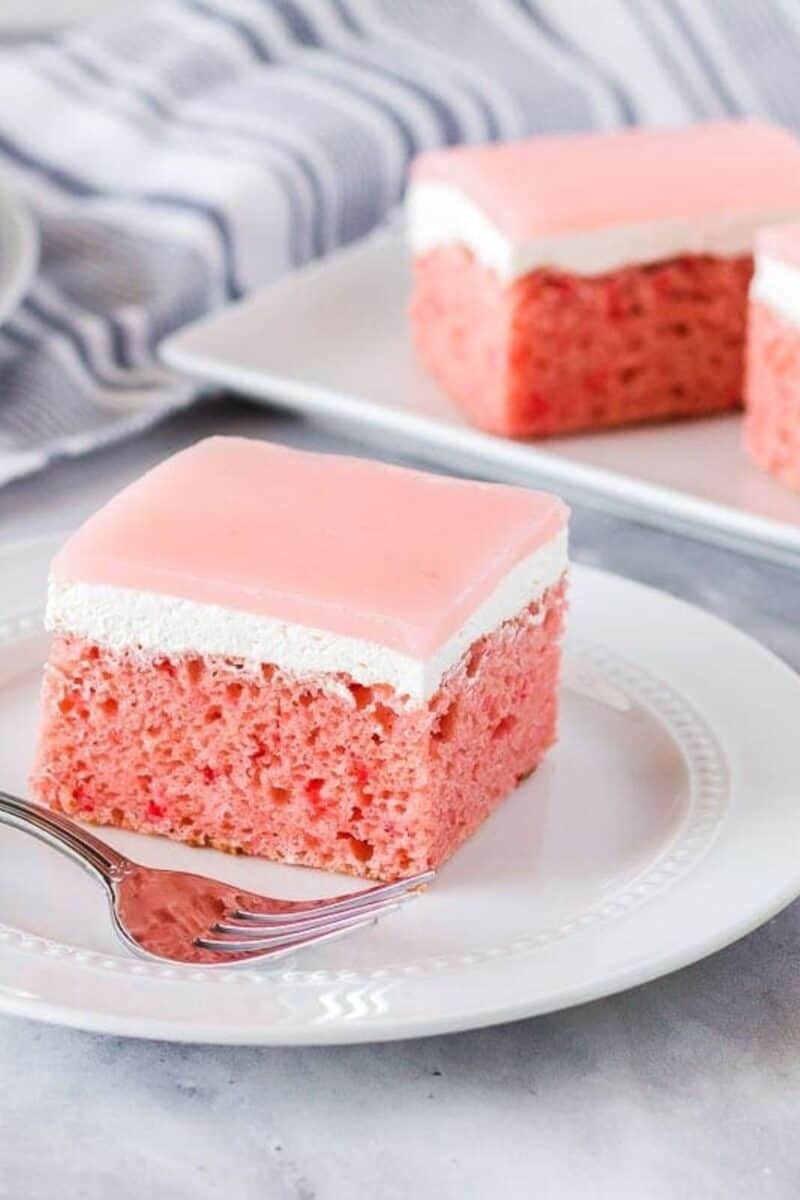 A pink guava cake with a layer of white frosting on top, cut into a square piece, presented on a white plate with a fork alongside.