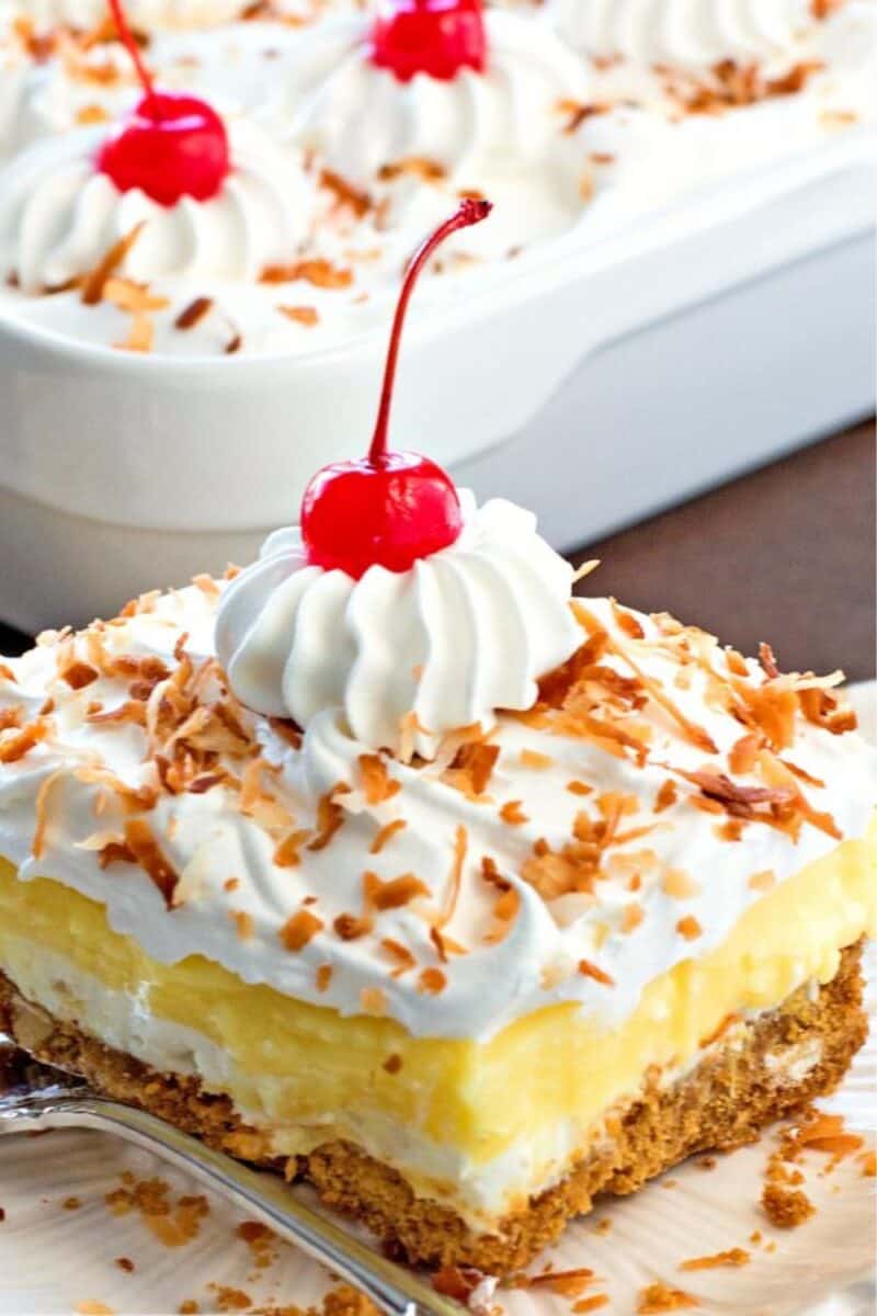 Image of a creamy dessert with a graham cracker crust, topped with a layer of bright yellow custard, whipped cream, toasted coconut, and a maraschino cherry.