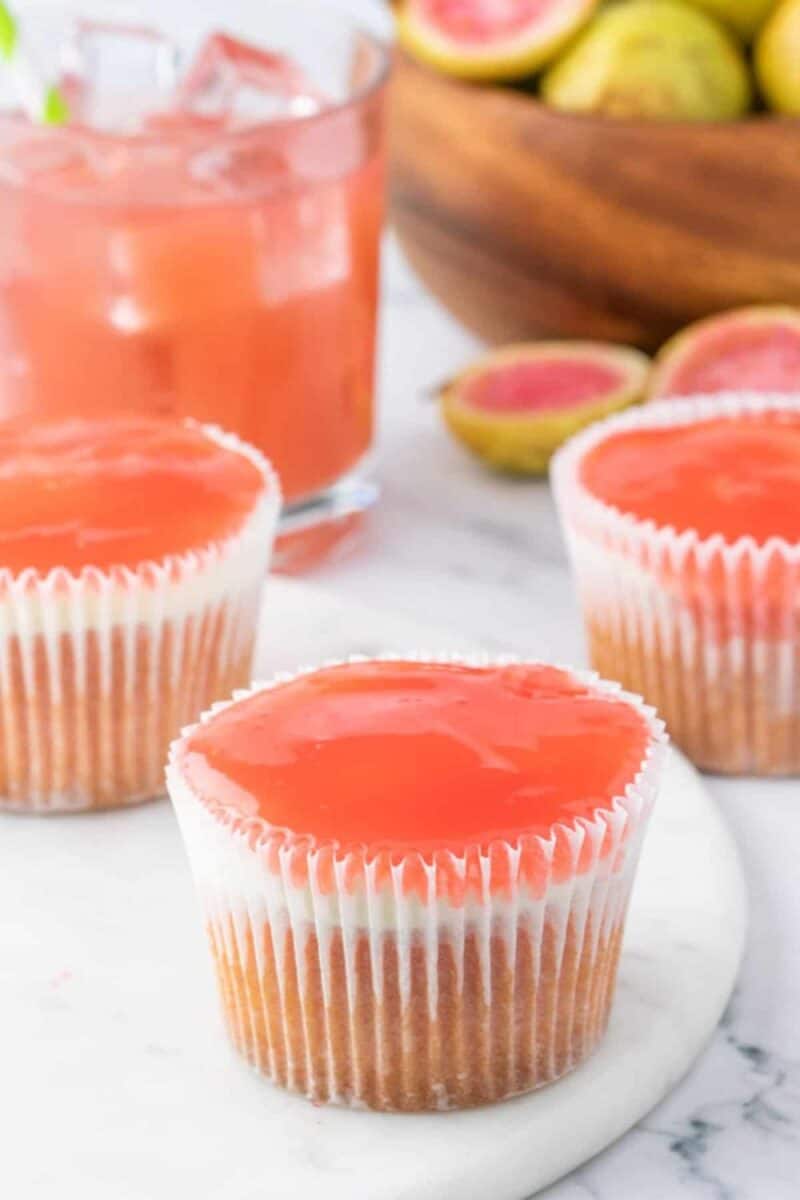 Cupcakes with a white base and a shiny, vibrant pink guava-flavored topping, presented on a marble surface with tropical fruits in the background, giving a refreshing and sweet look.