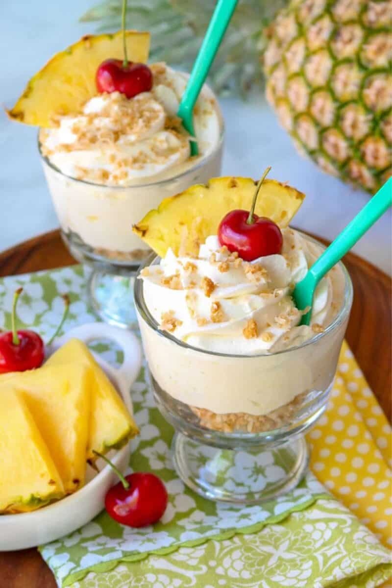 Two glasses of a layered tropical dessert, featuring whipped cream and crushed nuts, garnished with a slice of pineapple and a cherry, ready to be enjoyed with a straw.