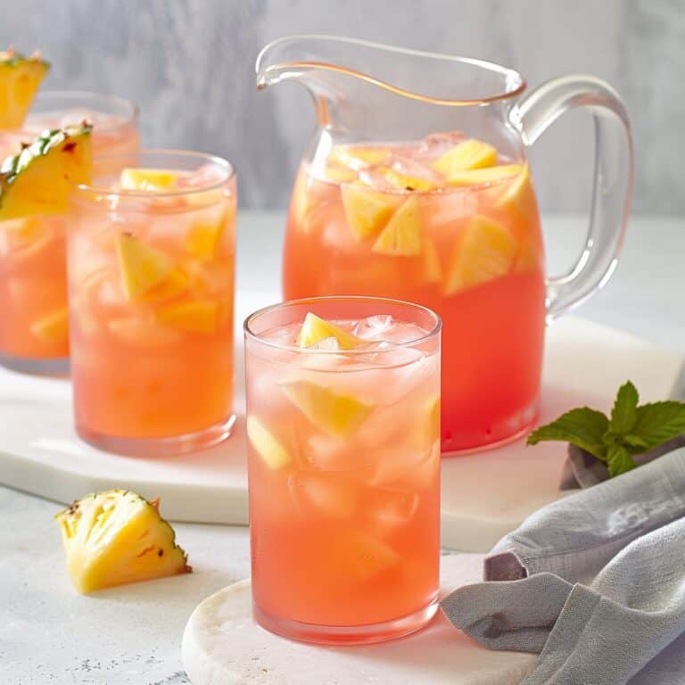 A vibrant pitcher filled with Pink Pineapple Lemonade sits on a white countertop, accompanied by glasses of the same refreshing drink, each garnished with a slice of pineapple and a twist of lemon, embodying a perfect summertime refreshment.