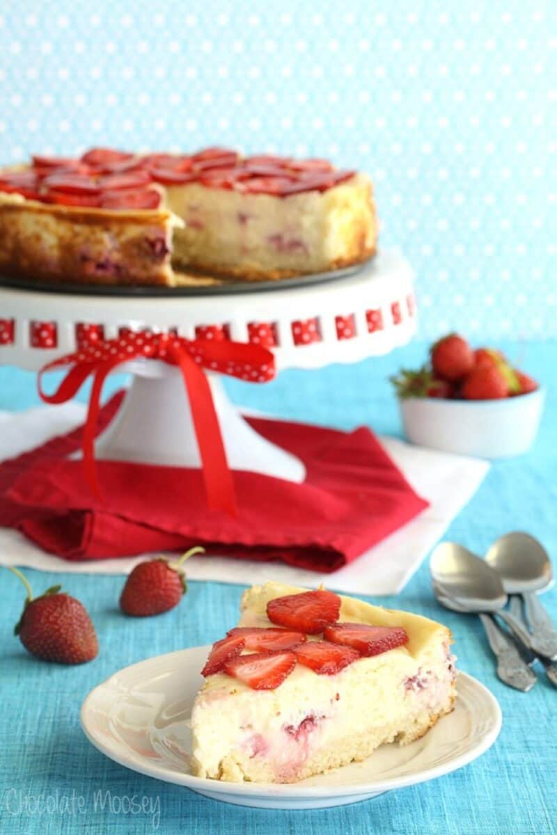 A festive strawberry shortcake cheesecake with a golden-brown crust, adorned with sliced strawberries on top, sits on a white cake stand with red ribbon details. In the foreground, a matching slice is plated, ready to be enjoyed.