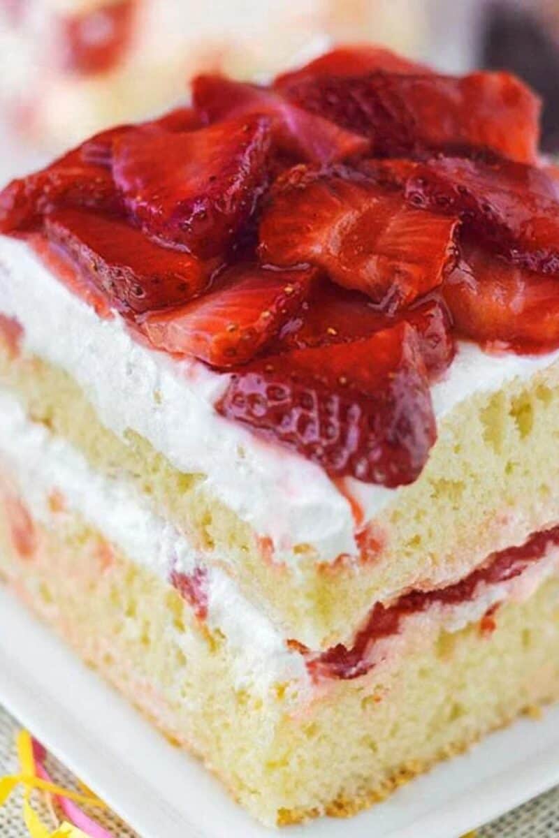 A slice of strawberry shortcake with layers of whipped cream and strawberries on a moist vanilla cake, presented on a plate with a festive ribbon.