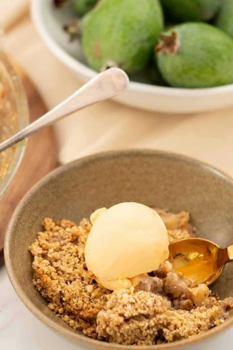 A scoop of feijoa ice cream atop a crumbly feijoa crumble dessert in a ceramic bowl, with whole feijoas and a spoon beside it.