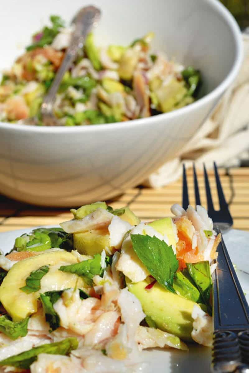A bowl of fresh feijoa salad with a mix of smoked fish, avocado slices, and herbs, presented on a bamboo mat with a fork on the side