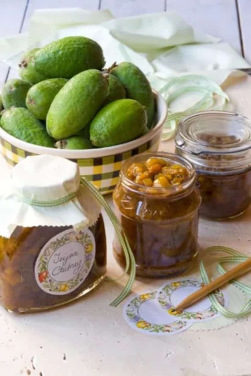 Homemade feijoa chutney in glass jars with checkered yellow and green ribbons, beside a bowl of fresh feijoas and parchment paper, ready for gifting or serving.