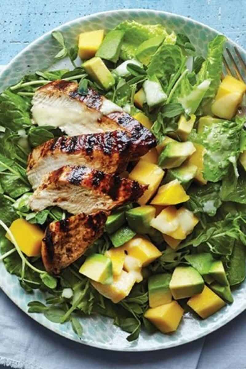 A fresh, vibrant salad featuring grilled chicken slices, chunks of mango and avocado, atop a bed of crisp greens on a decorative blue plate