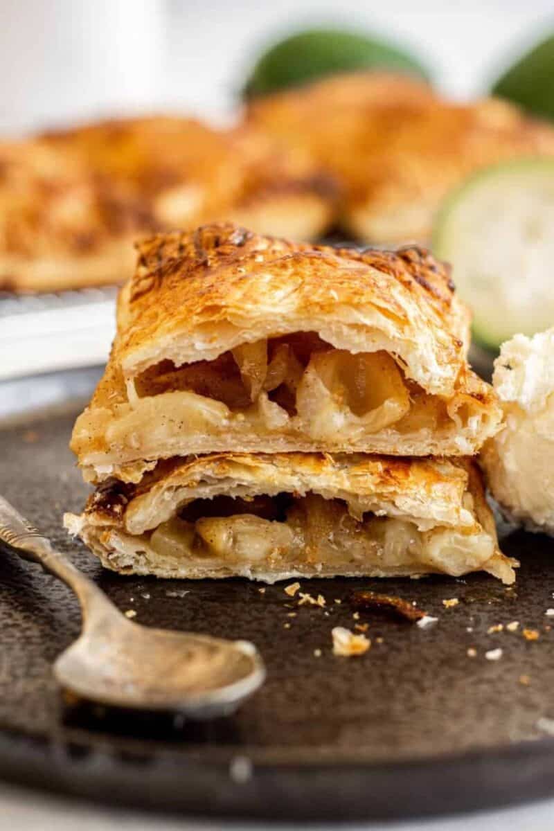 A golden-brown, flaky pastry with layers visible in the cross-section, filled with soft, caramelized feijoa slices, on a dark plate with a vintage spoon.