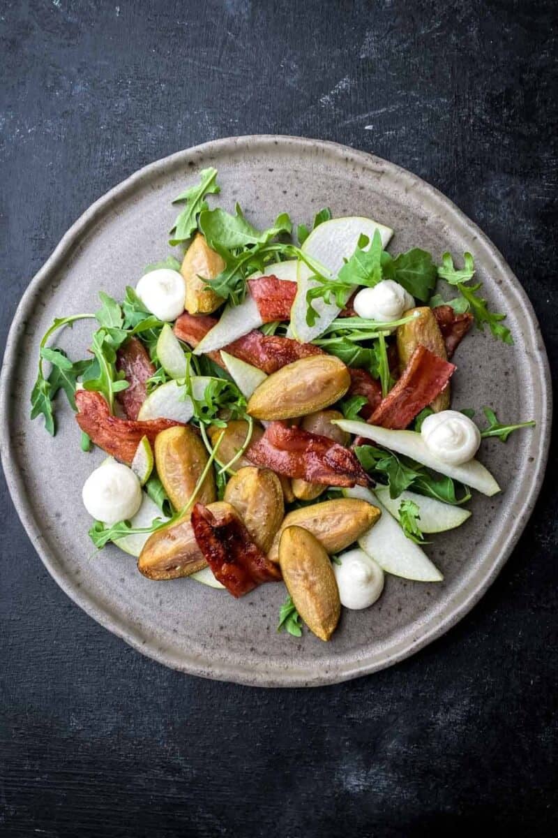 Artfully plated salad with roasted feijoas, streaky bacon, fresh mozzarella balls, and pear slices on a bed of arugula, served on a textured gray plate.
