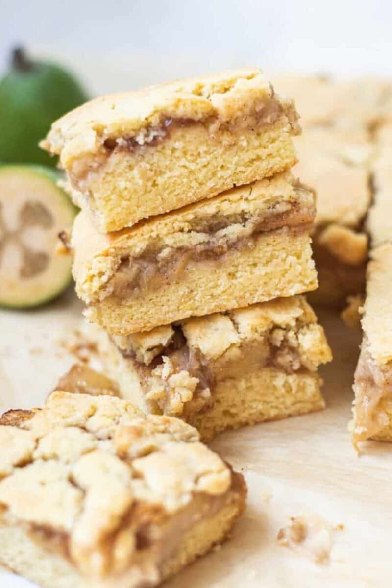 Stacked feijoa shortcake bars on parchment paper, showing layers of feijoa fruit filling, with a feijoa cut in half in the backdrop.